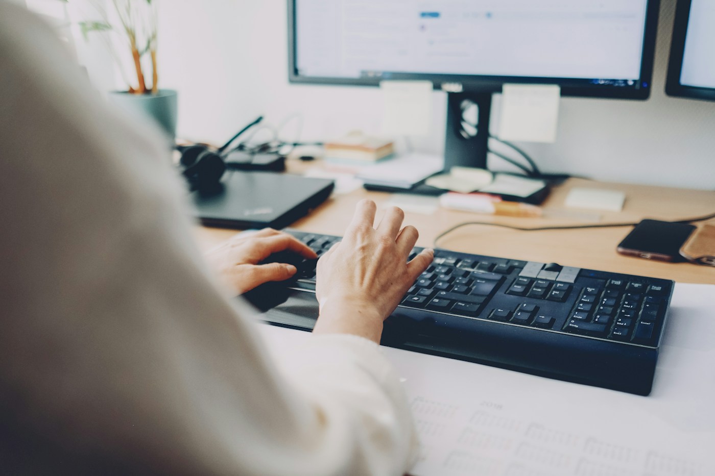 A person uses a keyboard at a desk with a computer and phone